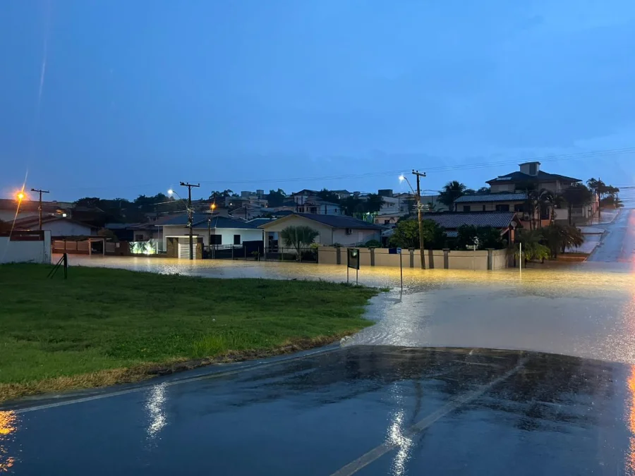 Chuva intensa alaga ruas e atinge casas em Cocal do Sul Fotografia de uma rua residencial em Cocal do Sul sob condições de alagamento ao anoitecer. A água barrenta cobre totalmente o asfalto, atingindo o nível das calçadas e muros das casas. As luzes dos postes de iluminação pública refletem na superfície da água. Ao fundo, veem-se casas em terrenos mais elevados e o céu em tons de azul escuro e cinza, indicando tempo nublado e chuvoso.