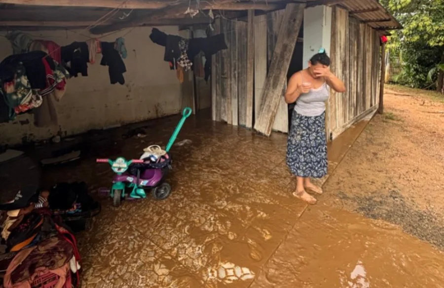 Fotografia colorida mostra uma mulher em um momento de desolação após a enxurrada em Cocal do Sul. Ela veste uma blusa cinza e saia estampada, segurando um copo e cobrindo o rosto com a mão em sinal de abatimento. O chão da varanda de sua casa de madeira está completamente coberto por uma espessa camada de lama marrom. Ao fundo, veem-se roupas penduradas em um varal improvisado e um triciclo infantil roxo e verde cercado pelos detritos trazidos pela inundação.