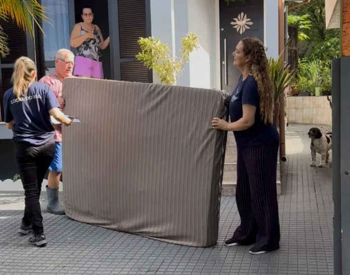 Cocal do Sul mobiliza doações para vítimas das chuvas Fotografia mostra duas mulheres e um homem transportando um colchão listrado em frente a uma residência. Uma das profissionais veste uma camiseta azul-marinho com a identificação "Cocal do Sul" estampada nas costas, evidenciando o trabalho das equipes municipais durante a coleta de doações para as famílias atingidas pelas chuvas no município.
