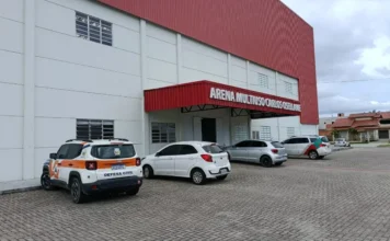 Cocal do Sul realiza simulado de Defesa Civil neste domingo Fachada da Arena Multiuso Carlos Osellame em Cocal do Sul, um prédio de paredes brancas com detalhe superior em metal vermelho. Em primeiro plano, no pátio de pavimentação intertravada, destaca-se um veículo utilitário branco com a identificação visual da Defesa Civil em laranja e preto. O céu está nublado, compondo o cenário onde será realizado o simulado de gestão de desastres.