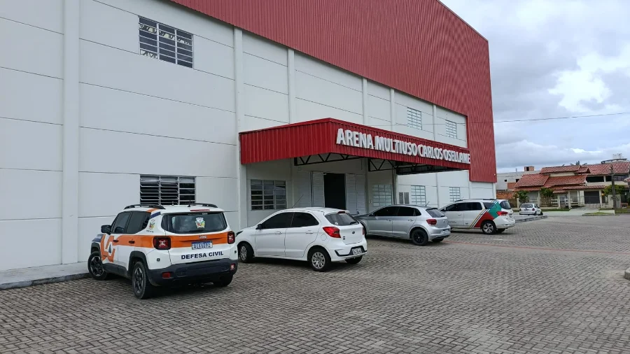 Fachada da Arena Multiuso Carlos Osellame em Cocal do Sul, um prédio de paredes brancas com detalhe superior em metal vermelho. Em primeiro plano, no pátio de pavimentação intertravada, destaca-se um veículo utilitário branco com a identificação visual da Defesa Civil em laranja e preto. O céu está nublado, compondo o cenário onde será realizado o simulado de gestão de desastres.