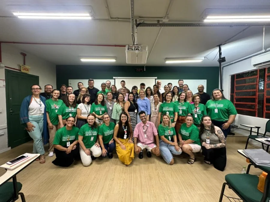 Fotografia em grupo reúne dezenas de professores e gestores do Colégio Unesc em uma sala de aula durante a formação continuada. Os profissionais estão sorridentes, alguns em pé e outros agachados à frente, muitos utilizando uma camiseta verde padronizada do colégio. O ambiente escolar é iluminado e conta com lousa e projetor ao fundo, simbolizando o momento de integração e planejamento pedagógico.