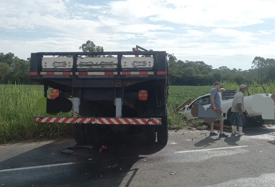 Cena do acidente na Rodovia Arino Antônio Cândido em Içara, mostrando a traseira do caminhão Ford Cargo parado na pista e a picape Saveiro branca fora da estrada, em uma área de vegetação, com duas pessoas em pé próximas a ela.