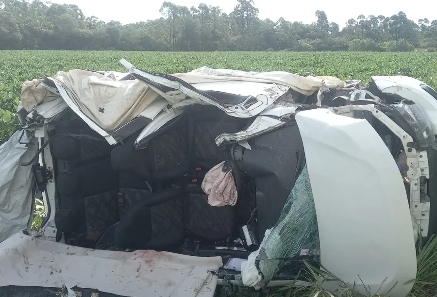 Destroços do veículo Fiat Mobi branco após o acidente em Içara, capotado em meio à vegetação. O teto do carro foi completamente removido pelas equipes de resgate do Corpo de Bombeiros para a extração das vítimas que estavam presas às ferragens.