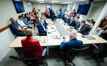 Comissão da Alesc aprova projeto sobre ICMS no agronegócio A imagem mostra a Comissão de Finanças e Tributação da Assembleia Legislativa de Santa Catarina (Alesc) em reunião para debater e votar o Projeto de Lei 68/2026 sobre o Tratamento Tributário Diferenciado (TTD) para o agronegócio. Vários deputados e membros da comissão estão sentados ao redor de uma grande mesa em formato de U, analisando documentos. O deputado Marcos Vieira (PSDB), relator da proposta, está entre eles. Um público composto por servidores, representantes do setor e imprensa acompanha a sessão ao fundo, alguns em pé e outros sentados. A sala é moderna, com painéis de TV e boa iluminação.