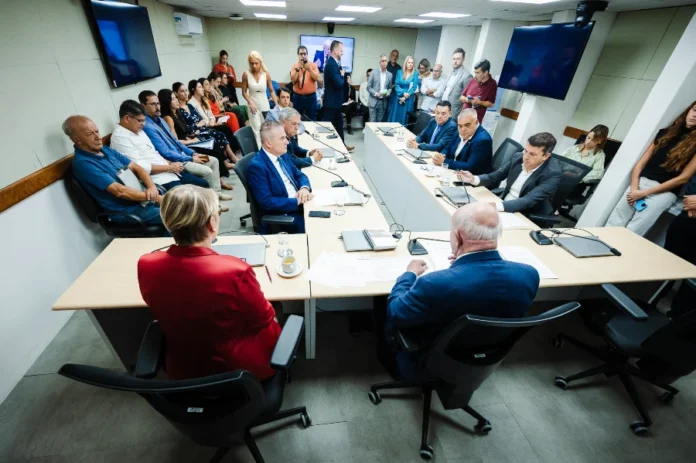 Comissão da Alesc aprova projeto sobre ICMS no agronegócio A imagem mostra a Comissão de Finanças e Tributação da Assembleia Legislativa de Santa Catarina (Alesc) em reunião para debater e votar o Projeto de Lei 68/2026 sobre o Tratamento Tributário Diferenciado (TTD) para o agronegócio. Vários deputados e membros da comissão estão sentados ao redor de uma grande mesa em formato de U, analisando documentos. O deputado Marcos Vieira (PSDB), relator da proposta, está entre eles. Um público composto por servidores, representantes do setor e imprensa acompanha a sessão ao fundo, alguns em pé e outros sentados. A sala é moderna, com painéis de TV e boa iluminação.