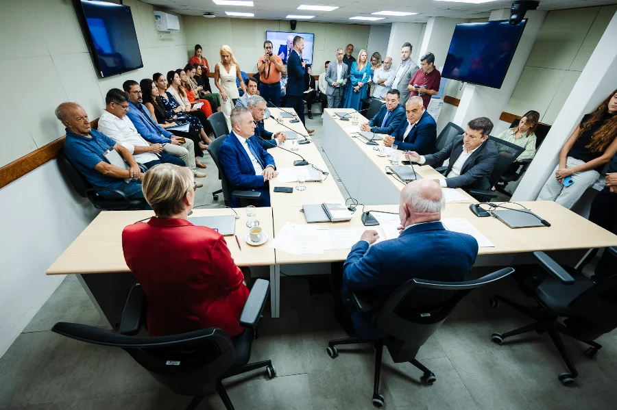 A imagem mostra a Comissão de Finanças e Tributação da Assembleia Legislativa de Santa Catarina (Alesc) em reunião para debater e votar o Projeto de Lei 68/2026 sobre o Tratamento Tributário Diferenciado (TTD) para o agronegócio. Vários deputados e membros da comissão estão sentados ao redor de uma grande mesa em formato de U, analisando documentos. O deputado Marcos Vieira (PSDB), relator da proposta, está entre eles. Um público composto por servidores, representantes do setor e imprensa acompanha a sessão ao fundo, alguns em pé e outros sentados. A sala é moderna, com painéis de TV e boa iluminação.