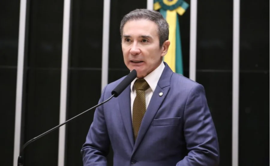 Fotografia do deputado federal Duda Ramos (MDB-RR) falando ao microfone no plenário da Câmara dos Deputados. Ele veste um terno azul, gravata dourada e está posicionado à frente de uma bancada preta com o mastro da bandeira do Brasil ao fundo.