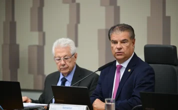 Congresso adia votação de acordo Mercosul-União Europeia O senador Nelsinho Trad, vice-presidente da Representação Brasileira no Parlamento do Mercosul, aparece em primeiro plano falando ao microfone durante a reunião que discutiu o acordo comercial com a União Europeia. Ele está sentado à mesa, veste terno azul-marinho e gravata rosa, com um notebook e sua identificação oficial à frente. Ao fundo, à esquerda, outro parlamentar de cabelos brancos observa documentos em seu próprio computador em um ambiente institucional.