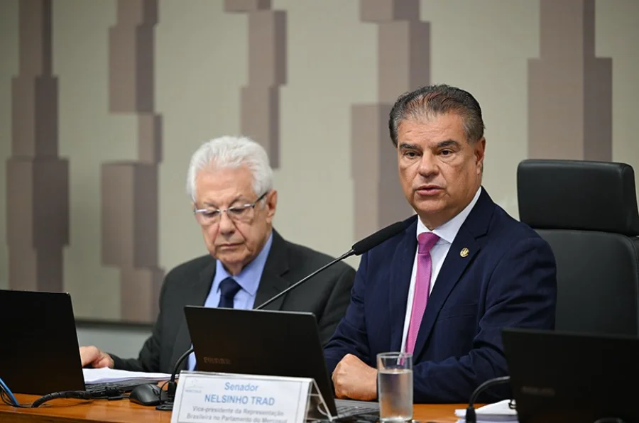 O senador Nelsinho Trad, vice-presidente da Representação Brasileira no Parlamento do Mercosul, aparece em primeiro plano falando ao microfone durante a reunião que discutiu o acordo comercial com a União Europeia. Ele está sentado à mesa, veste terno azul-marinho e gravata rosa, com um notebook e sua identificação oficial à frente. Ao fundo, à esquerda, outro parlamentar de cabelos brancos observa documentos em seu próprio computador em um ambiente institucional.