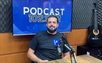 Construtora de Araranguá entrega casas em apenas 30 dias O empresário Diego Natal Mateus, proprietário da Construtora Casas Paraíso, aparece sentado nos estúdios da Rádio Amorim FM. Ele é um homem de cabelos escuros e barba, veste uma camiseta preta e sorri para a câmera. À sua frente, há um microfone profissional de rádio e, ao fundo, uma tela digital exibe o logotipo "PODCAST 102.9 FM". O ambiente é composto por painéis acústicos e um violão posicionado à direita.