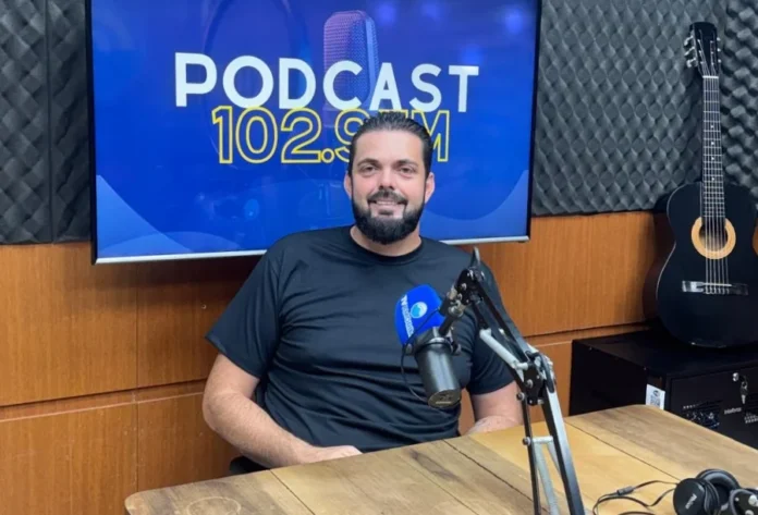 Construtora de Araranguá entrega casas em apenas 30 dias O empresário Diego Natal Mateus, proprietário da Construtora Casas Paraíso, aparece sentado nos estúdios da Rádio Amorim FM. Ele é um homem de cabelos escuros e barba, veste uma camiseta preta e sorri para a câmera. À sua frente, há um microfone profissional de rádio e, ao fundo, uma tela digital exibe o logotipo "PODCAST 102.9 FM". O ambiente é composto por painéis acústicos e um violão posicionado à direita.