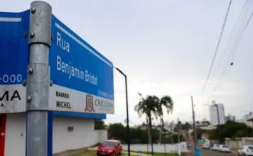 Criciúma altera tráfego na Benjamin Bristot para segurança Placa azul de identificação urbana em primeiro plano com os dizeres "Rua Benjamin Bristot" e "Bairro Michel", exibindo o brasão da Prefeitura de Criciúma. Ao fundo, em perspectiva, vê-se a via pavimentada com lajotas, veículos estacionados e árvores, ilustrando o trecho que recebeu a alteração para sentido único de circulação.