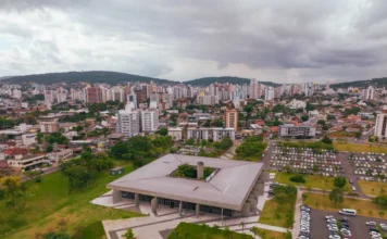 Criciúma atinge nota máxima em transparência da CGU Vista aérea do Paço Municipal de Criciúma, um edifício de arquitetura quadrada com um pátio central verde, cercado por um amplo estacionamento e áreas gramadas. Ao fundo, observa-se a densa paisagem urbana com prédios e morros sob um céu nublado, representando o centro administrativo que obteve a nota máxima em transparência.