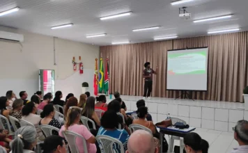 Criciúma capacita professores em relações étnico-raciais Foto do auditório durante a capacitação de professores da rede municipal de Criciúma. No palco, um palestrante negro aponta para uma tela de projeção que exibe um slide com o título "POR QUE PLANEJAR EM ERER?". Uma plateia composta por dezenas de educadores sentados em cadeiras brancas assiste atentamente à apresentação. Ao fundo, estão as bandeiras do Brasil, de Santa Catarina e do município.