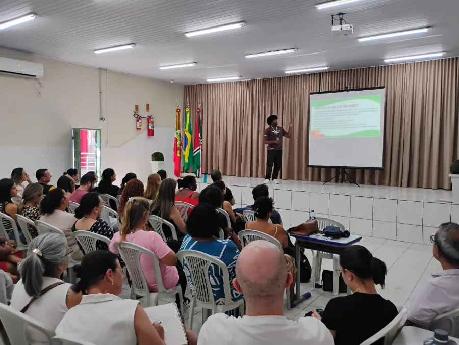 Foto do auditório durante a capacitação de professores da rede municipal de Criciúma. No palco, um palestrante negro aponta para uma tela de projeção que exibe um slide com o título 