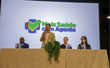 Criciúma forma 116 novos agentes para a saúde municipal O prefeito de Criciúma, Vagner Espindola, aparece de pé ao centro de uma mesa de autoridades, discursando ao microfone durante a formatura dos agentes de saúde. Ao fundo, um telão exibe o logotipo do programa "Mais Saúde com Agente". Ladeando o prefeito, estão sentados outros três representantes da gestão municipal e da saúde. A imagem registra o evento realizado no Auditório Ruy Hülse, na Unesc.