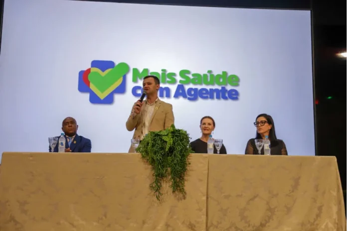 Criciúma forma 116 novos agentes para a saúde municipal O prefeito de Criciúma, Vagner Espindola, aparece de pé ao centro de uma mesa de autoridades, discursando ao microfone durante a formatura dos agentes de saúde. Ao fundo, um telão exibe o logotipo do programa "Mais Saúde com Agente". Ladeando o prefeito, estão sentados outros três representantes da gestão municipal e da saúde. A imagem registra o evento realizado no Auditório Ruy Hülse, na Unesc.