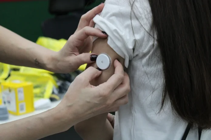 Criciúma oferece sensores de glicose gratuitos para crianças Fotografia em close-up detalha o momento em que um profissional de saúde aplica um sensor de monitoramento contínuo de glicose, de formato circular e cor branca, na parte posterior do braço de uma criança. O dispositivo substitui as frequentes picadas no dedo, facilitando o controle do diabetes tipo 1 sem causar dor. No plano de fundo, é possível observar materiais de suporte clínico em um ambiente de atendimento de saúde.