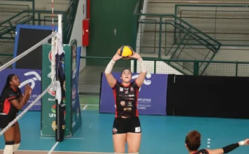 Criciúma recebe Ceará no Ginásio da Unesc nesta terça Uma jogadora da equipe de voleibol Mampituba/FME Criciúma/Radar/Unesc, vestindo uniforme preto e vermelho com patrocinadores, está em quadra realizando um toque com as duas mãos acima da cabeça durante uma partida. Ela está perto da rede, e outras jogadoras podem ser vistas ao redor. O ginásio possui arquibancadas verdes e banners de patrocinadores ao fundo, incluindo a marca da Unesc.