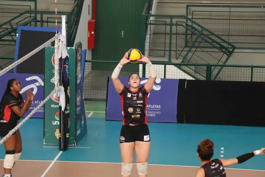 Uma jogadora da equipe de voleibol Mampituba/FME Criciúma/Radar/Unesc, vestindo uniforme preto e vermelho com patrocinadores, está em quadra realizando um toque com as duas mãos acima da cabeça durante uma partida. Ela está perto da rede, e outras jogadoras podem ser vistas ao redor. O ginásio possui arquibancadas verdes e banners de patrocinadores ao fundo, incluindo a marca da Unesc.