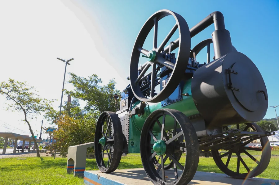 A imagem mostra o locomóvel centenário de 1914, um equipamento industrial a vapor com grandes rodas de ferro e uma chaminé, após seu processo de restauração. Ele está pintado nas cores preta e verde e posicionado sobre uma base de concreto ao ar livre, sob um céu azul ensolarado na Praça da Chaminé, em Criciúma.