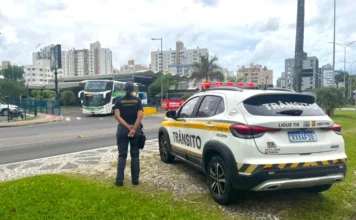 Criciúma sedia 1º Encontro Estadual de Agentes de Trânsito Fotografia em plano médio de uma agente de trânsito de Criciúma, vista de costas, utilizando uniforme escuro com a inscrição "Trânsito" em amarelo. Ela está parada ao lado de uma viatura oficial branca e amarela, modelo SUV, estacionada em um canteiro gramado. Ao fundo, observa-se o cenário urbano de Criciúma, com um ônibus em movimento e edifícios sob um céu nublado.