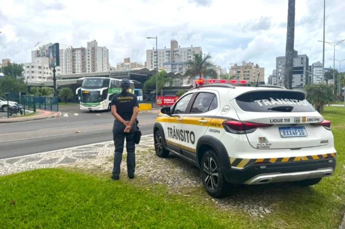Criciúma sedia 1º Encontro Estadual de Agentes de Trânsito Fotografia em plano médio de uma agente de trânsito de Criciúma, vista de costas, utilizando uniforme escuro com a inscrição "Trânsito" em amarelo. Ela está parada ao lado de uma viatura oficial branca e amarela, modelo SUV, estacionada em um canteiro gramado. Ao fundo, observa-se o cenário urbano de Criciúma, com um ônibus em movimento e edifícios sob um céu nublado.