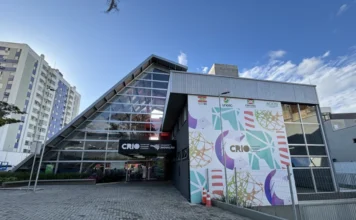 Criciúma sedia Meetup Startup SC e apresenta o MIDIHUB Vista frontal externa do Centro de Inovação de Criciúma (CRIO) durante o dia. O prédio possui uma arquitetura moderna com uma grande fachada de vidro em formato triangular à esquerda e uma parede cinza à direita decorada com grafismos coloridos e logotipos institucionais. O letreiro na entrada identifica o local como o hub de inovação da região, onde ocorrerá o Meetup Startup SC.