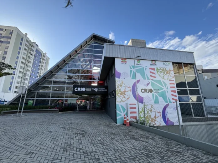 Criciúma sedia Meetup Startup SC e apresenta o MIDIHUB Vista frontal externa do Centro de Inovação de Criciúma (CRIO) durante o dia. O prédio possui uma arquitetura moderna com uma grande fachada de vidro em formato triangular à esquerda e uma parede cinza à direita decorada com grafismos coloridos e logotipos institucionais. O letreiro na entrada identifica o local como o hub de inovação da região, onde ocorrerá o Meetup Startup SC.