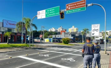 Criciúma testa área de espera para motos em semáforos Agentes de trânsito de Criciúma observam a nova área de espera experimental para motocicletas, sinalizada com pintura no asfalto e placa vertical, no cruzamento da Avenida Centenário. A imagem ilustra a medida de segurança viária implantada pela DTT.