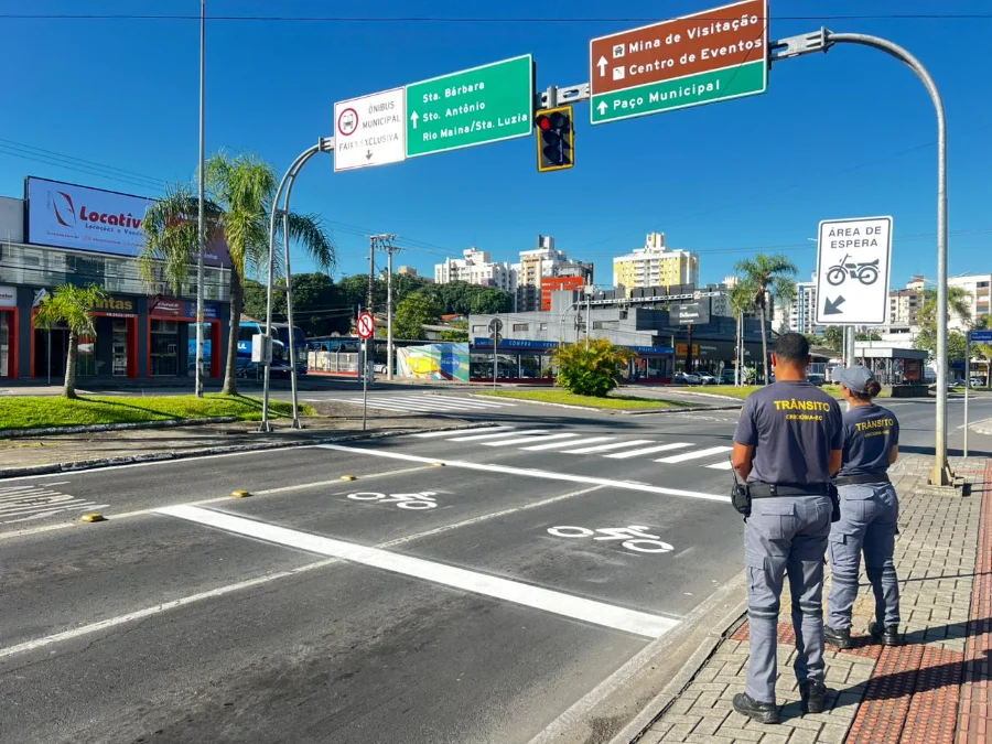 Agentes de trânsito de Criciúma observam a nova área de espera experimental para motocicletas, sinalizada com pintura no asfalto e placa vertical, no cruzamento da Avenida Centenário. A imagem ilustra a medida de segurança viária implantada pela DTT.