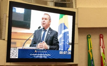 Deputado com esclerose usa IA para discursar em SC Fotografia de um grande monitor digital instalado no plenário da Assembleia Legislativa de Santa Catarina (Alesc). A tela exibe, em primeiro plano, o deputado Maurício Eskudlark (PL) vestido de terno e gravata, discursando atrás de uma tribuna com microfones. Na parte inferior do monitor, consta a inscrição "10ª Sessão Plenária Ordinária – 25 de fevereiro de 2026", acompanhada do brasão do estado. Ao lado direito do equipamento, é possível observar as bandeiras físicas do Brasil e de Santa Catarina posicionadas no ambiente.