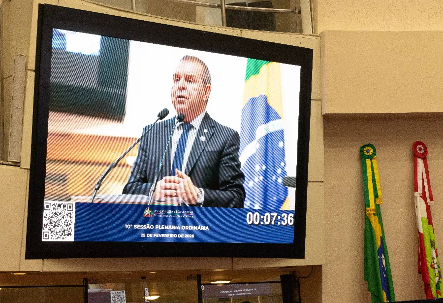 Fotografia de um grande monitor digital instalado no plenário da Assembleia Legislativa de Santa Catarina (Alesc). A tela exibe, em primeiro plano, o deputado Maurício Eskudlark (PL) vestido de terno e gravata, discursando atrás de uma tribuna com microfones. Na parte inferior do monitor, consta a inscrição 
