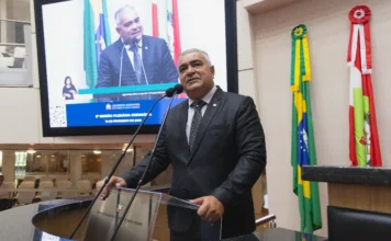 Deputado processa escola de samba por enredo sobre Lula A imagem mostra o deputado estadual Sargento Lima, um homem de pele clara e cabelos curtos grisalhos, vestindo terno preto, camisa branca e gravata escura. Ele está posicionado atrás de um púlpito de acrílico transparente com o brasão do Estado de Santa Catarina, falando ao microfone na tribuna da Assembleia Legislativa (Alesc). Ao fundo, um grande monitor exibe a transmissão ao vivo de seu próprio discurso, acompanhado por uma intérprete de Libras no canto da tela. À direita do parlamentar, as bandeiras do Brasil e de Santa Catarina estão dispostas verticalmente.
