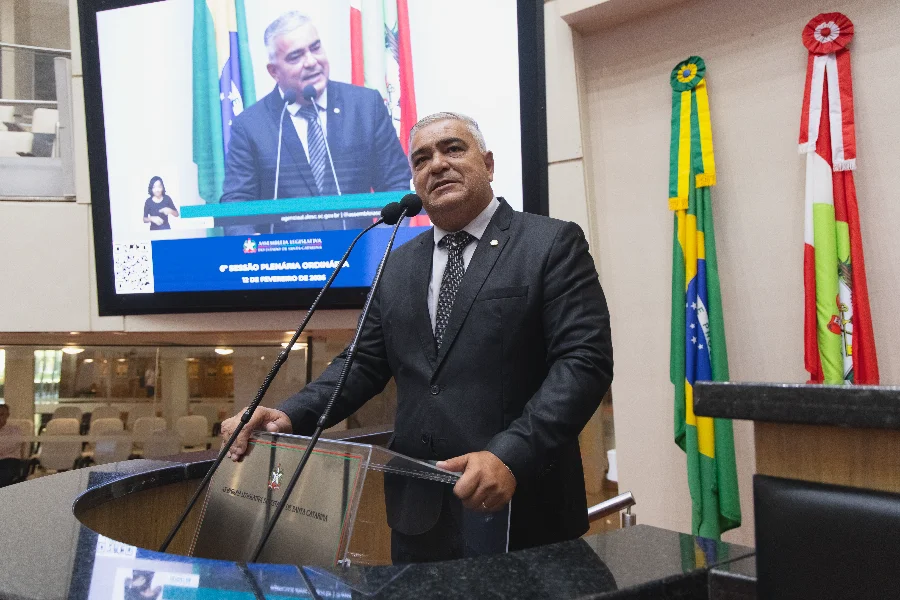 A imagem mostra o deputado estadual Sargento Lima, um homem de pele clara e cabelos curtos grisalhos, vestindo terno preto, camisa branca e gravata escura. Ele está posicionado atrás de um púlpito de acrílico transparente com o brasão do Estado de Santa Catarina, falando ao microfone na tribuna da Assembleia Legislativa (Alesc). Ao fundo, um grande monitor exibe a transmissão ao vivo de seu próprio discurso, acompanhado por uma intérprete de Libras no canto da tela. À direita do parlamentar, as bandeiras do Brasil e de Santa Catarina estão dispostas verticalmente.