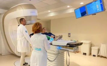 Dia Mundial do Câncer: SC foca em diagnóstico precoce A imagem mostra um paciente deitado em uma maca móvel sendo posicionado dentro de um grande equipamento de radioterapia ou diagnóstico por imagem de alta tecnologia. Dois profissionais de saúde, um homem e uma mulher, ambos vestindo jalecos brancos, acompanham o procedimento. O homem está de pé ao lado da máquina, enquanto a mulher segura um controle remoto. A cena ilustra o investimento em tecnologia de ponta e a estrutura de atendimento especializado disponíveis na rede de saúde de Santa Catarina para o diagnóstico e tratamento do câncer, conforme abordado na matéria.
