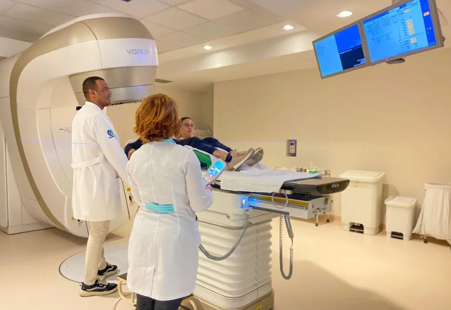 A imagem mostra um paciente deitado em uma maca móvel sendo posicionado dentro de um grande equipamento de radioterapia ou diagnóstico por imagem de alta tecnologia. Dois profissionais de saúde, um homem e uma mulher, ambos vestindo jalecos brancos, acompanham o procedimento. O homem está de pé ao lado da máquina, enquanto a mulher segura um controle remoto. A cena ilustra o investimento em tecnologia de ponta e a estrutura de atendimento especializado disponíveis na rede de saúde de Santa Catarina para o diagnóstico e tratamento do câncer, conforme abordado na matéria.