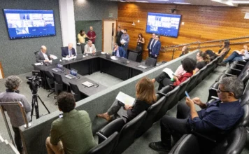 Doenças crônicas: lei de SC fixa cadastro estadual em 2026 Fotografia de uma reunião da comissão da ALESC, onde vários deputados e assessores estão sentados ao redor de uma longa mesa branca em formato de U. Eles utilizam laptops e microfones. Ao fundo, uma parede de vidro exibe o brasão da Assembleia Legislativa de Santa Catarina e, em uma tela grande na parede lateral, há uma videoconferência com múltiplos participantes. O ambiente é bem iluminado e formal.
