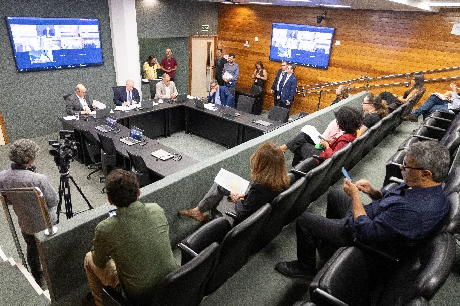 Fotografia de uma reunião da comissão da ALESC, onde vários deputados e assessores estão sentados ao redor de uma longa mesa branca em formato de U. Eles utilizam laptops e microfones. Ao fundo, uma parede de vidro exibe o brasão da Assembleia Legislativa de Santa Catarina e, em uma tela grande na parede lateral, há uma videoconferência com múltiplos participantes. O ambiente é bem iluminado e formal.