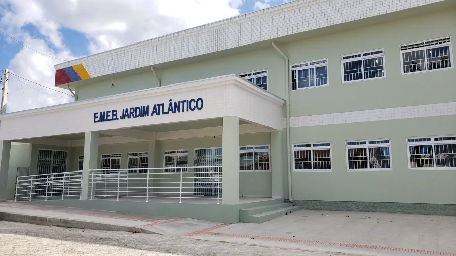 Fotografia da fachada da Escola Municipal de Educação Básica Jardim Atlântico, em Balneário Arroio do Silva. O prédio de dois andares apresenta pintura em tons de verde claro e branco, com rampas de acessibilidade na entrada e uma placa azul com o nome da escola em destaque. O local é o ponto de atendimento noturno para as matrículas da EJA.