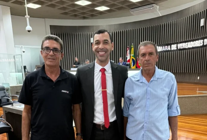 ETA da Lagoa do Caverá recebe nome de Ésio João Roque Foto no plenário da Câmara de Vereadores mostrando o vereador Márcio Mano, autor do projeto, de terno e gravata vermelha ao centro, sorrindo ao lado de dois homens, familiares do homenageado Ésio João Roque, após a aprovação da lei que dá nome à Estação de Tratamento de Água.