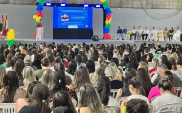 Educação: Encontro reúne mil profissionais em Santa Rosa Vista da parte de trás de uma grande plateia composta majoritariamente por mulheres, profissionais da educação, sentadas em cadeiras brancas. Elas estão voltadas para um palco decorado com arcos de balões coloridos (azul, amarelo e vermelho). No centro do palco, um telão azul exibe a foto do palestrante Dado Schneider e o título do evento sobre conflitos geracionais. À direita do palco, autoridades locais estão sentadas em linha, acompanhando a cerimônia no pavilhão do Parque de Eventos da Polvilhana.