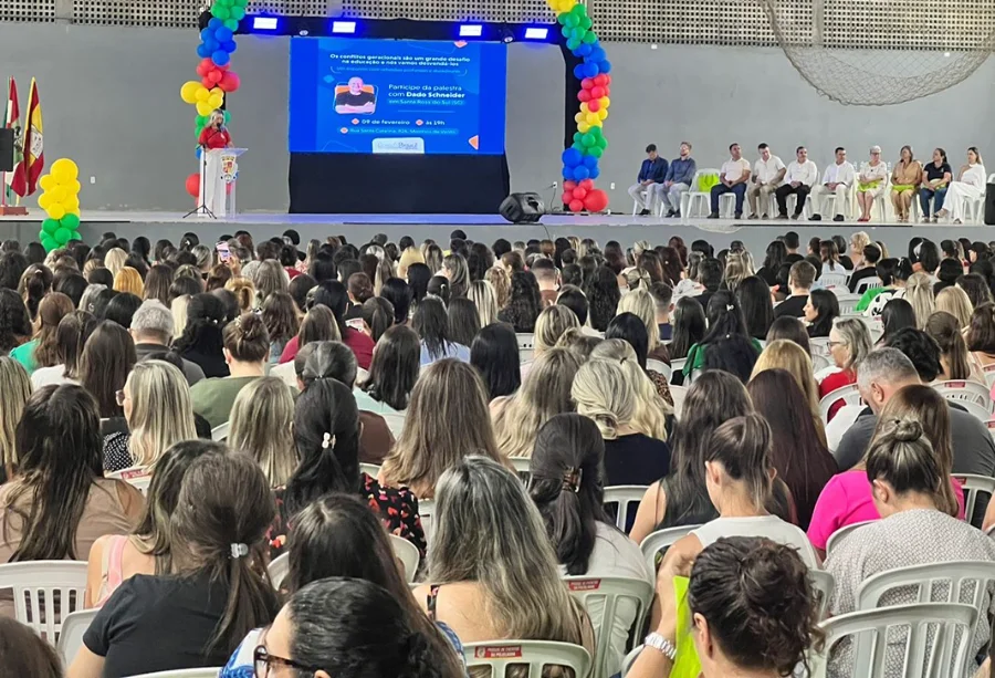 Vista da parte de trás de uma grande plateia composta majoritariamente por mulheres, profissionais da educação, sentadas em cadeiras brancas. Elas estão voltadas para um palco decorado com arcos de balões coloridos (azul, amarelo e vermelho). No centro do palco, um telão azul exibe a foto do palestrante Dado Schneider e o título do evento sobre conflitos geracionais. À direita do palco, autoridades locais estão sentadas em linha, acompanhando a cerimônia no pavilhão do Parque de Eventos da Polvilhana.