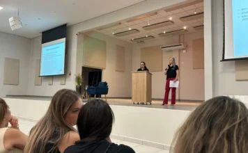 Educação de Araranguá alinha RH para o ano letivo 2026 Duas mulheres no palco do Centro Multiuso de Araranguá realizam uma apresentação para servidores da Educação. Uma das palestrantes está atrás de um púlpito de madeira, enquanto a outra fala ao microfone segurando papéis. Ao fundo, telas de projeção exibem o título "Integração para Servidores" com o brasão do município. Em primeiro plano, é possível ver parte da plateia acompanhando a formação.