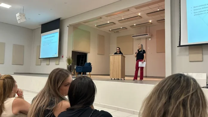 Educação de Araranguá alinha RH para o ano letivo 2026 Duas mulheres no palco do Centro Multiuso de Araranguá realizam uma apresentação para servidores da Educação. Uma das palestrantes está atrás de um púlpito de madeira, enquanto a outra fala ao microfone segurando papéis. Ao fundo, telas de projeção exibem o título "Integração para Servidores" com o brasão do município. Em primeiro plano, é possível ver parte da plateia acompanhando a formação.