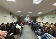 Entidades de Araranguá debatem Reforma Tributária Fotografia do auditório da ACIVA, em Araranguá, preenchido por dezenas de pessoas sentadas em fileiras de cadeiras marrons. O público, composto por homens e mulheres, assiste atentamente à palestra sobre a Reforma Tributária. Ao fundo da sala, centralizada na parede branca, está a logomarca verde da ACIVA. Os palestrantes Jandir Pereira e Rodrigo Nolla aparecem sentados à frente, conduzindo o debate. Nas paredes laterais, destacam-se frases motivacionais sobre empreendedorismo em tons de azul e verde.