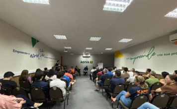 Entidades de Araranguá debatem Reforma Tributária Fotografia do auditório da ACIVA, em Araranguá, preenchido por dezenas de pessoas sentadas em fileiras de cadeiras marrons. O público, composto por homens e mulheres, assiste atentamente à palestra sobre a Reforma Tributária. Ao fundo da sala, centralizada na parede branca, está a logomarca verde da ACIVA. Os palestrantes Jandir Pereira e Rodrigo Nolla aparecem sentados à frente, conduzindo o debate. Nas paredes laterais, destacam-se frases motivacionais sobre empreendedorismo em tons de azul e verde.