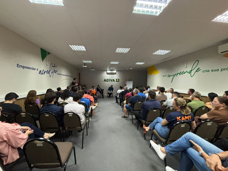 Fotografia do auditório da ACIVA, em Araranguá, preenchido por dezenas de pessoas sentadas em fileiras de cadeiras marrons. O público, composto por homens e mulheres, assiste atentamente à palestra sobre a Reforma Tributária. Ao fundo da sala, centralizada na parede branca, está a logomarca verde da ACIVA. Os palestrantes Jandir Pereira e Rodrigo Nolla aparecem sentados à frente, conduzindo o debate. Nas paredes laterais, destacam-se frases motivacionais sobre empreendedorismo em tons de azul e verde.