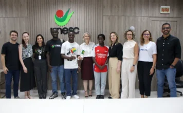Estudantes africanos ingressam na Unesc via programa federal Uma fotografia de grupo tirada na reitoria da Unesc, capturando a recepção oficial de três estudantes de Guiné-Bissau pelo Programa de Estudantes-Convênio de Graduação (PEC-G). No centro, os estudantes posam ao lado da reitora em exercício da Unesc, Gisele Silveira Coelho Lopes, que segura uma pequena bandeira do Brasil, enquanto um dos alunos segura a bandeira de Guiné-Bissau. Uma das estudantes usa uma camisa de futebol do Arsenal. A imagem inclui membros da equipe administrativa da Unesc e da coordenação do PEC-G, com o logotipo da universidade visível ao fundo em um painel de madeira.